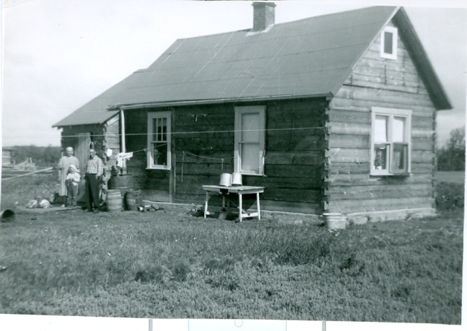Black and white photograph of a farmhouse with people standing in front of it, near barrels and a table